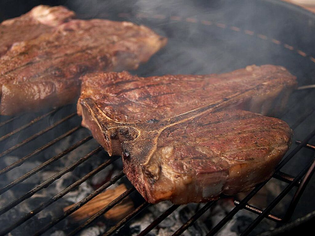 Overhead angled shot of two browned porterhouse steaks sizzling on a grill over coals, steam and smoke rising from them