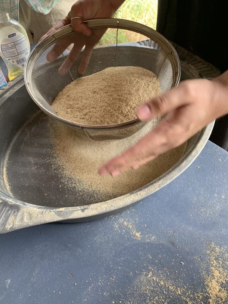 Scenes from an outdoor mesquite milling event, with a large mill on the back of a trailer parked in front of a home, participants in masks, and mesquite being sorted, roasted, milled, then sifted into a fine, pale sand-colored flour.