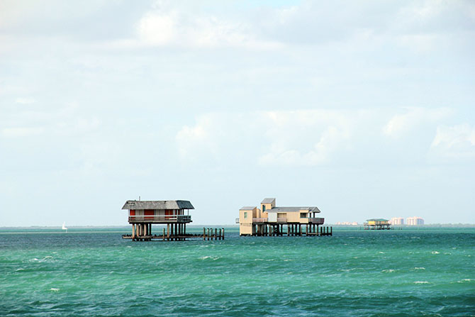 A photo of three colorful houses on tall wooden poles perched in bright green-blue waters. A shoreline is visible in the distance.