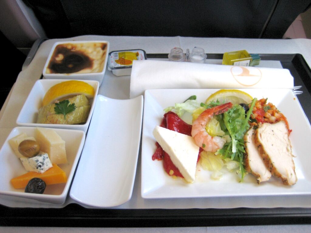 Photograph of a meal on a airplane tray featuring some slices of chicken breast, a shrimp, and a piece of cheese on salad, some small dishes to the side with fruit and other sides, and a tiny salt and pepper shaker at the top