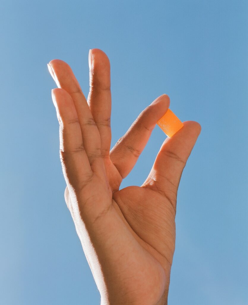 A hand holds up a single orange gummy between the index finger and thumb against the background of a bright blue sky