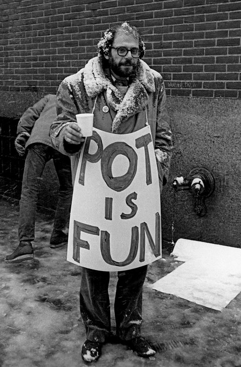A black and white photo of a bearded and bespectacled Allen Ginsberg with snow on his head and coat collar holding up a large sign that reads "POT IS FUN."