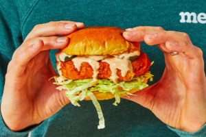 Side view of two hands holding a sandwich containing a crispy chicken cutlet, shredded cabbage, and a creamy sauce