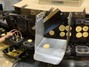 A yellow disk of cookie flies off a rotating grilling machine and into a metal catch pan to be folded.