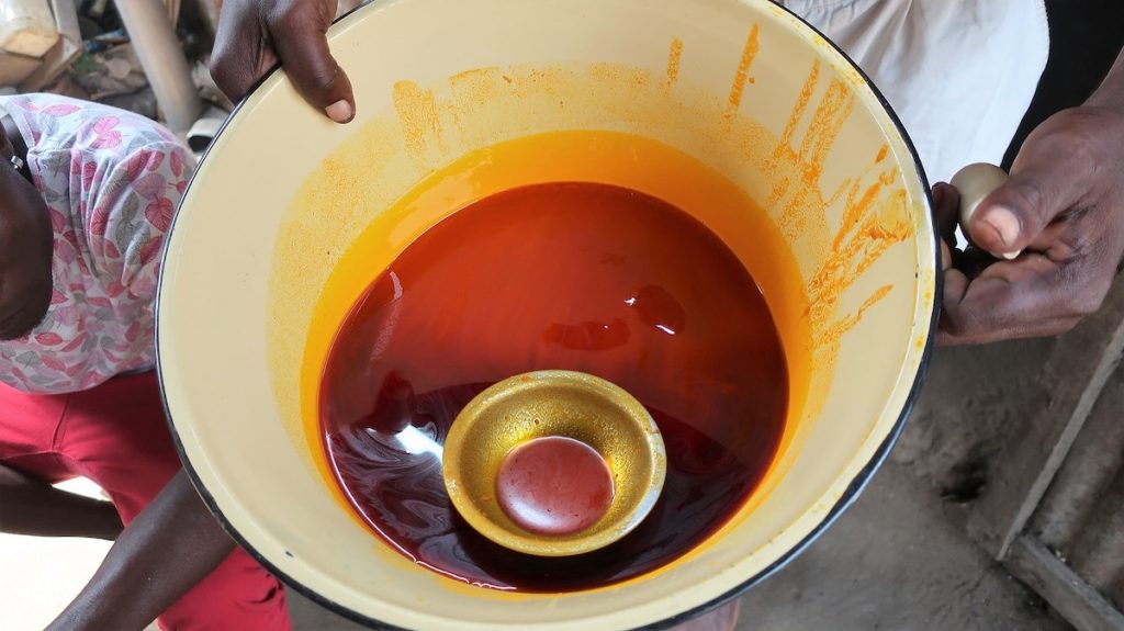 A brown hand holds a large white bowl beneath the camera. Inside, a deep reddish-orange liquid is visible. A small bowl for scooping floats on the surface.