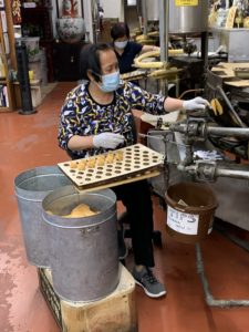 An older woman in a mask and colorful shirt sits at a tray punctured with holes, each of which holds the bright yellow fortune cookies she is folding as they fly off the machine.