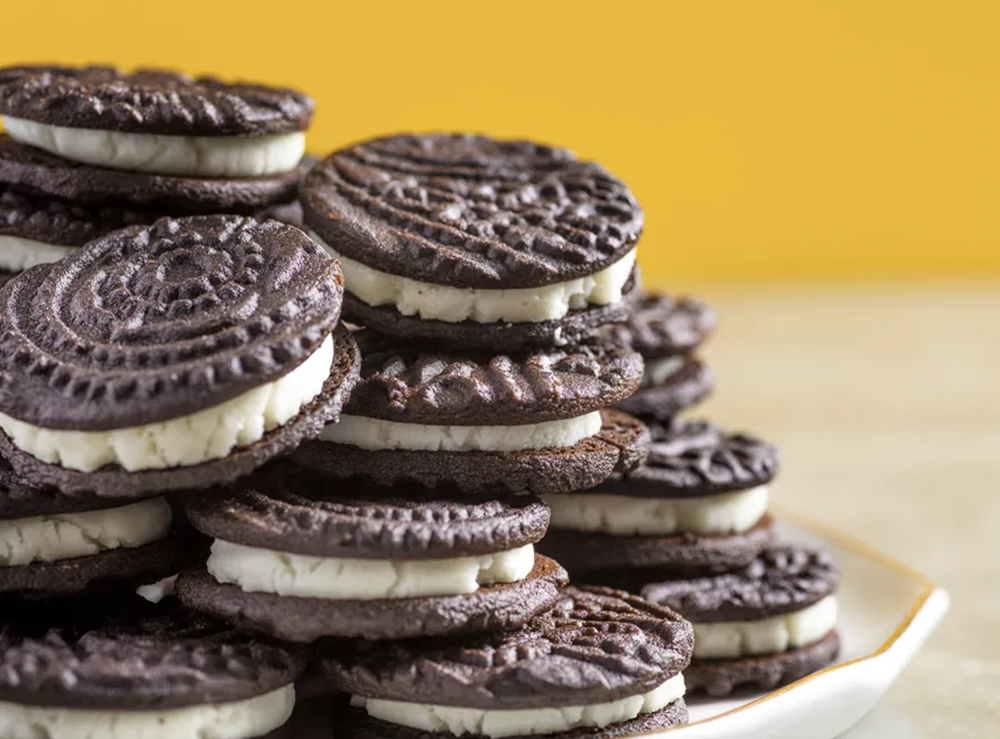 A pile of homemade dark chocolate sandwich cookies with elaborate swirls on the chocolate cookie and white cream centers, on a yellow background.